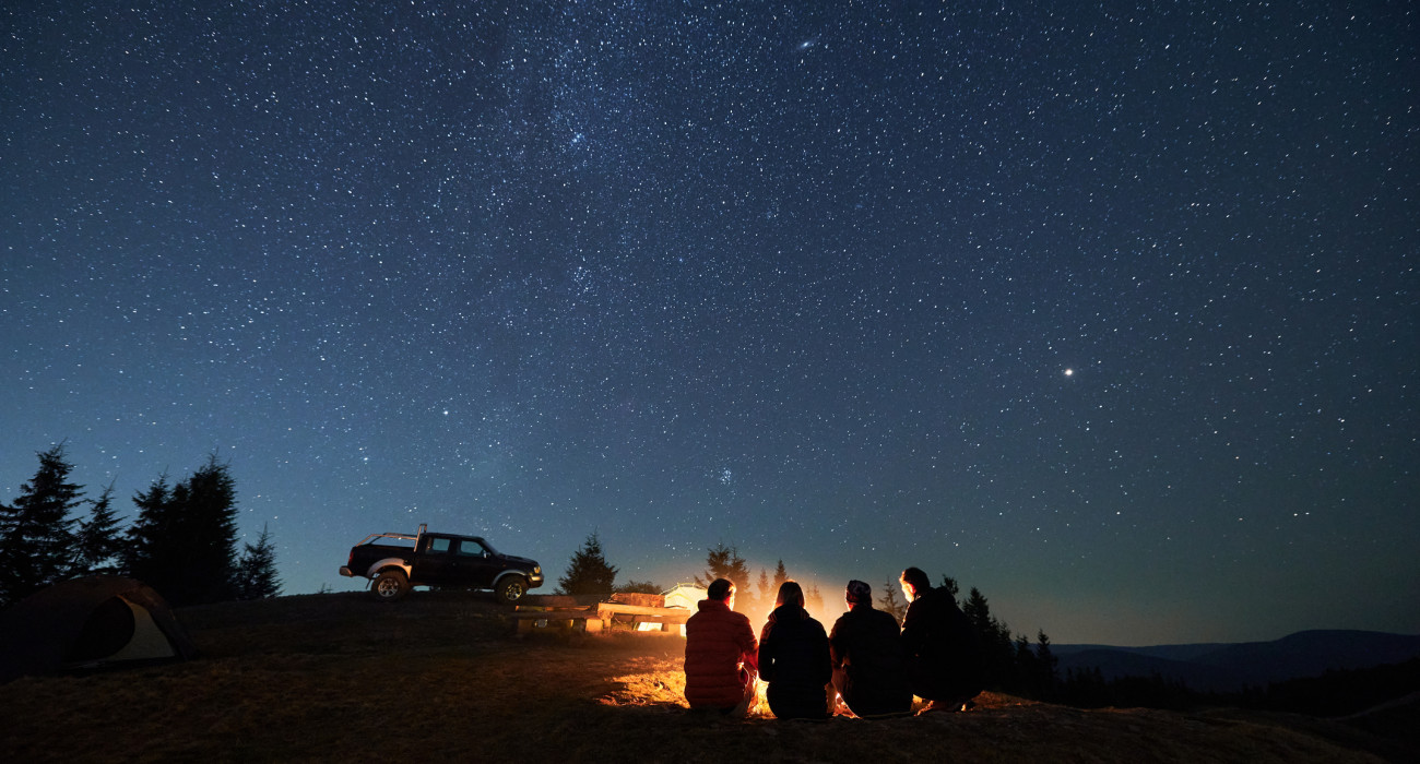 Evening starry sky over mountain valley with car and hikers near campfire. Group of travelers sitting near bonfire under majestic blue sky with stars. Concept of night camping, hiking and travelling.